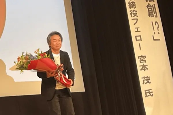 Shigeru Miyamoto holding a bouquet on stage next to a banner; source: https://note.com/hibiki_linasha/n/nd10546afe57a